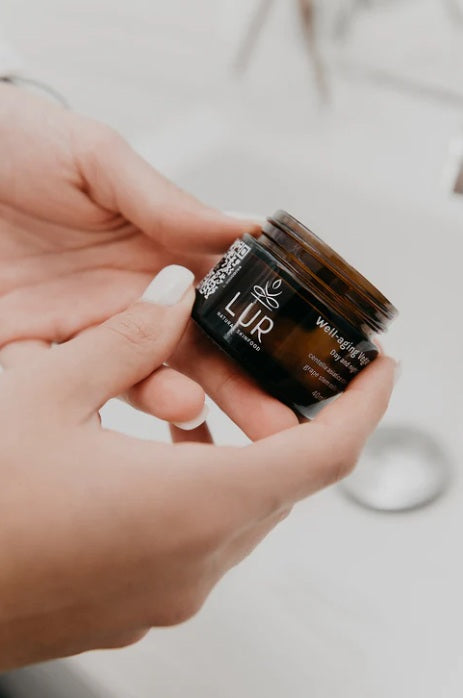 Handen die een amberkleurige pot met gezichtscrème vasthouden in een badkamer.