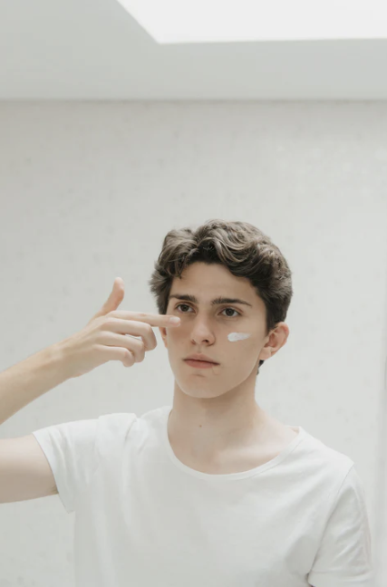 Jonge man in wit shirt brengt gezichtscrème aan op zijn wang in een lichte ruimte.