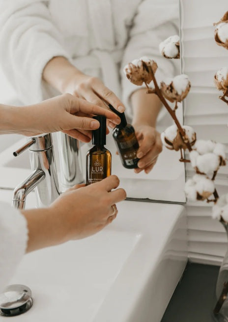 Persoon in witte badjas met glazen pipetflesje bij de wastafel in de badkamer, met katoenbloemen.
